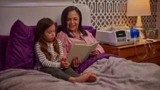 A women is reading to her granddaughter in her bed. The Homechoice Claria system sits on her nightstand